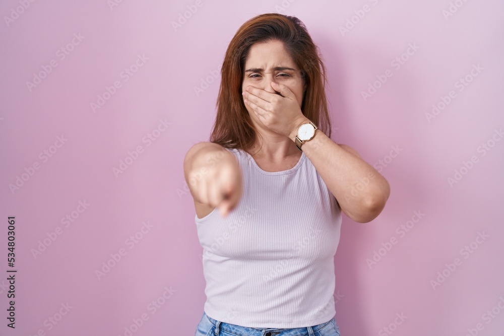 Brunette woman standing over pink background laughing at you, pointing finger to the camera with hand over mouth, shame expression Brunette woman standing over pink background laughing at you, pointing finger to the camera with hand over mouth, shame expression