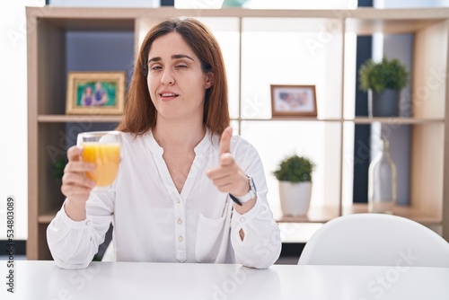 Brunette woman drinking glass of orange juice pointing fingers to camera with happy and funny face. good energy and vibes.