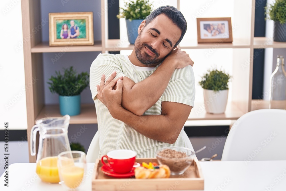 Hispanic man with beard eating breakfast hugging oneself happy and positive, smiling confident. self love and self care Hispanic man with beard eating breakfast hugging oneself happy and positive, smiling confident. self love and self care