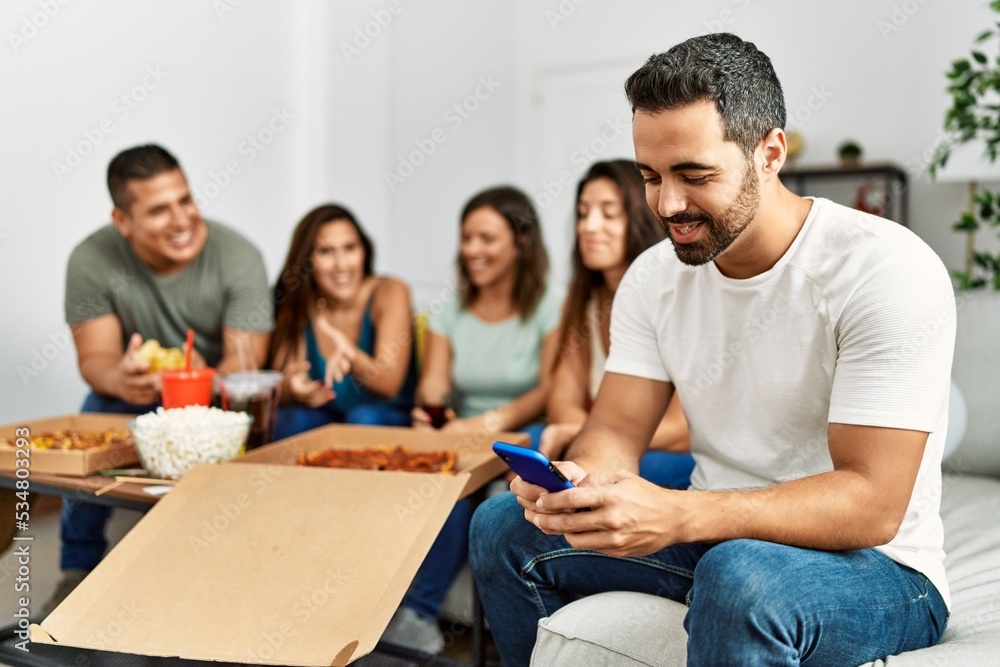 Fototapeta premium Group of young hispanic friends eating italian pizza sitting on the sofa. Man smiling happy and using smartphone at home.