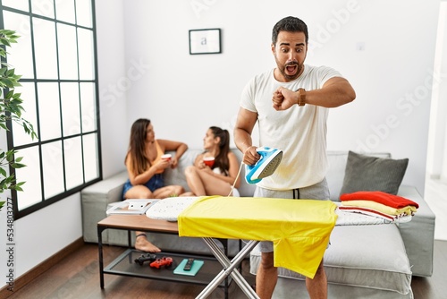 Young hispanic man ironing clothes at home looking at the watch time worried  afraid of getting late