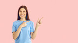 Happy beautiful young woman on pink background advertises new promotion or offers sale. Smiling friendly woman in blue t-shirt looking at camera and pointing with two index fingers to side.