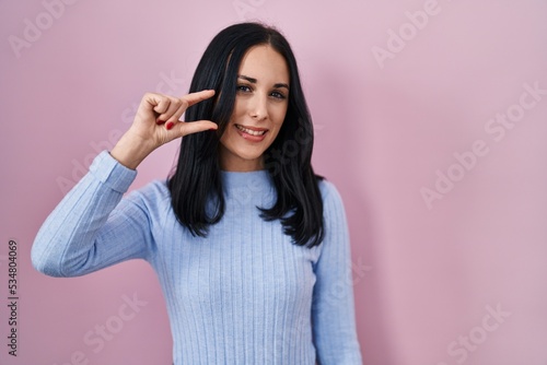 Hispanic woman standing over pink background smiling and confident gesturing with hand doing small size sign with fingers looking and the camera. measure concept.