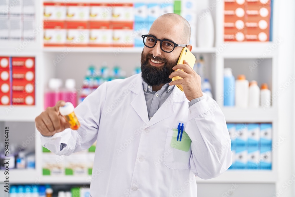 Young bald man pharmacist holding pills bottle talking on smartphone at pharmacy