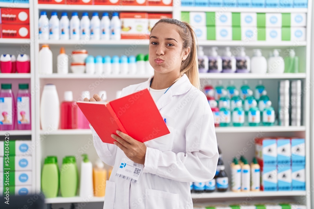 Young blonde woman working at pharmacy drugstore holding notebook puffing cheeks with funny face. mouth inflated with air, catching air.