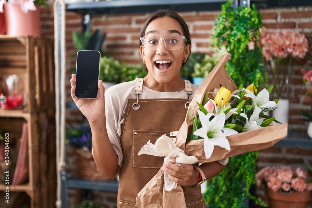 Young hispanic woman working at florist shop showing smartphone screen celebrating crazy and amazed for success with open eyes screaming excited.