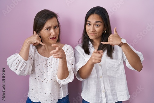 Hispanic mother and daughter together smiling doing talking on the telephone gesture and pointing to you. call me.