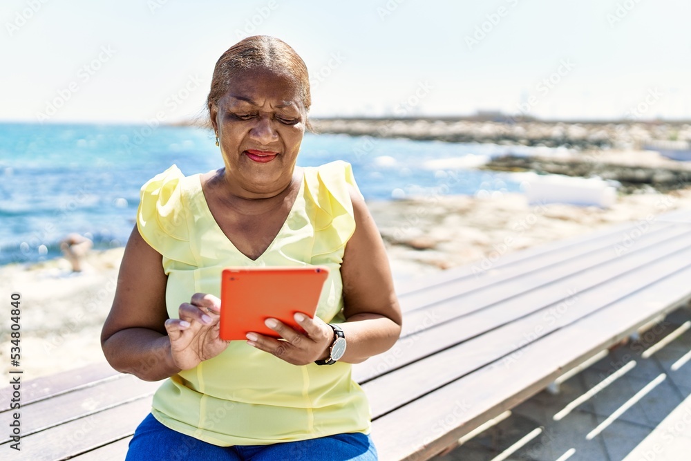 Senior african american woman using touchpad sitting on the bench at the beach.