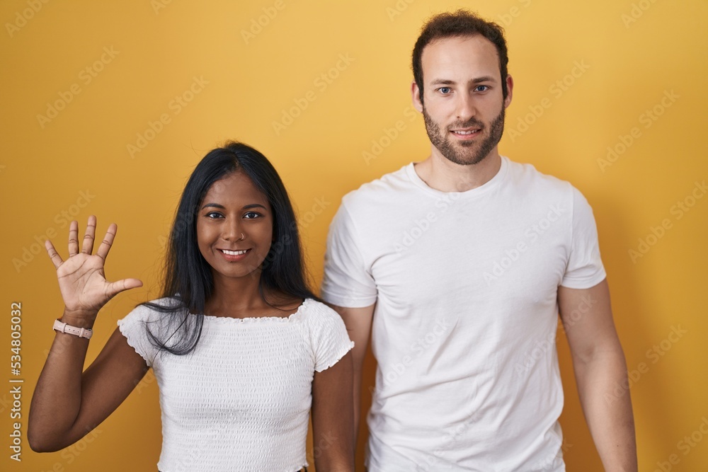 Interracial couple standing over yellow background showing and pointing up with fingers number five while smiling confident and happy. Interracial couple standing over yellow background showing and pointing up with fingers number five while smiling confident and happy.
