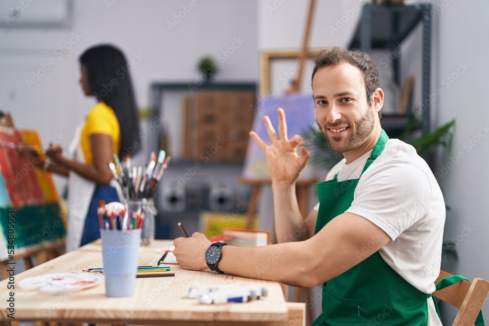 Fototapeta premium Hispanic man at painter studio doing ok sign with fingers, smiling friendly gesturing excellent symbol