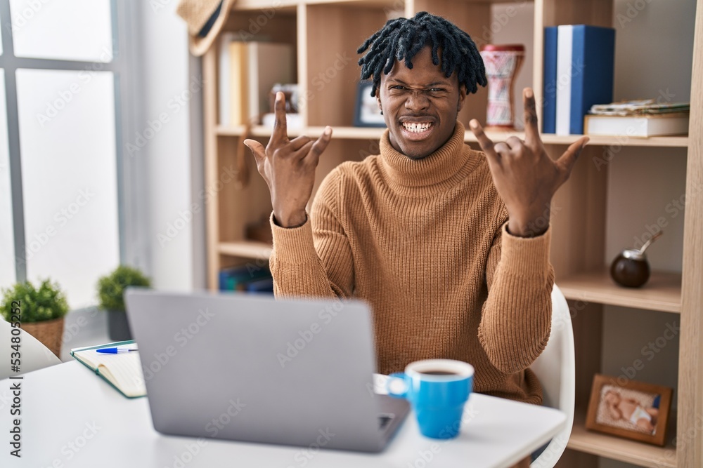 Young african man with dreadlocks working using computer laptop shouting with crazy expression doing rock symbol with hands up. music star. heavy concept. Young african man with dreadlocks working using computer laptop shouting with crazy expression doing rock symbol with hands up. music star. heavy concept.