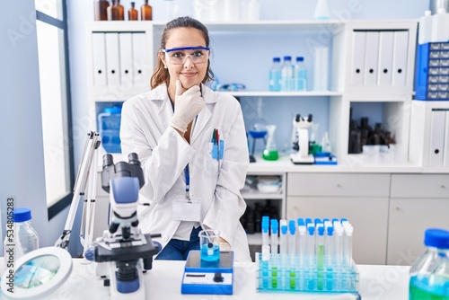 Young hispanic woman working at scientist laboratory looking confident at the camera with smile with crossed arms and hand raised on chin. thinking positive.
