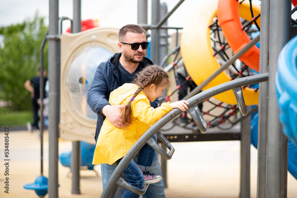 A girl 6-7 years old with her dad on the playground. She on the slide and has a fun. Dad helps the girl climb the stairs. Childhood. Time with family. Spring. City. A girl 6-7 years old with her dad on the playground. She on the slide and has a fun. Dad helps the girl climb the stairs. Childhood. Time with family. Spring. City.