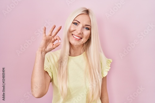 Caucasian woman standing over pink background smiling positive doing ok sign with hand and fingers. successful expression.