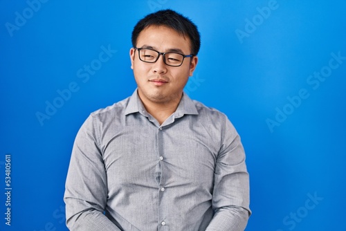 Young chinese man standing over blue background smiling looking to the side and staring away thinking.