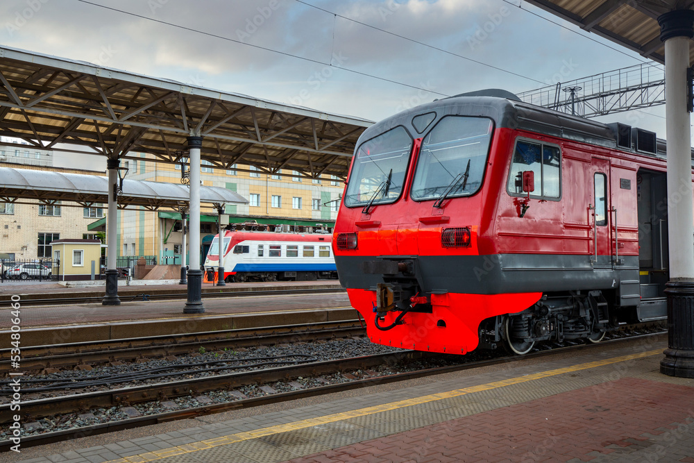 A bright red train stands on the platform of the railway station in the city. Passenger Transportation. A bright red train stands on the platform of the railway station in the city. Passenger Transportation.
