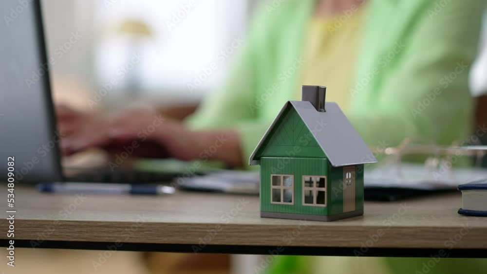 Close-up toy house with hands of blurred woman typing on laptop keyboard at background. Unrecognizable Caucasian real estate agent working online indoors in office. Slow motion