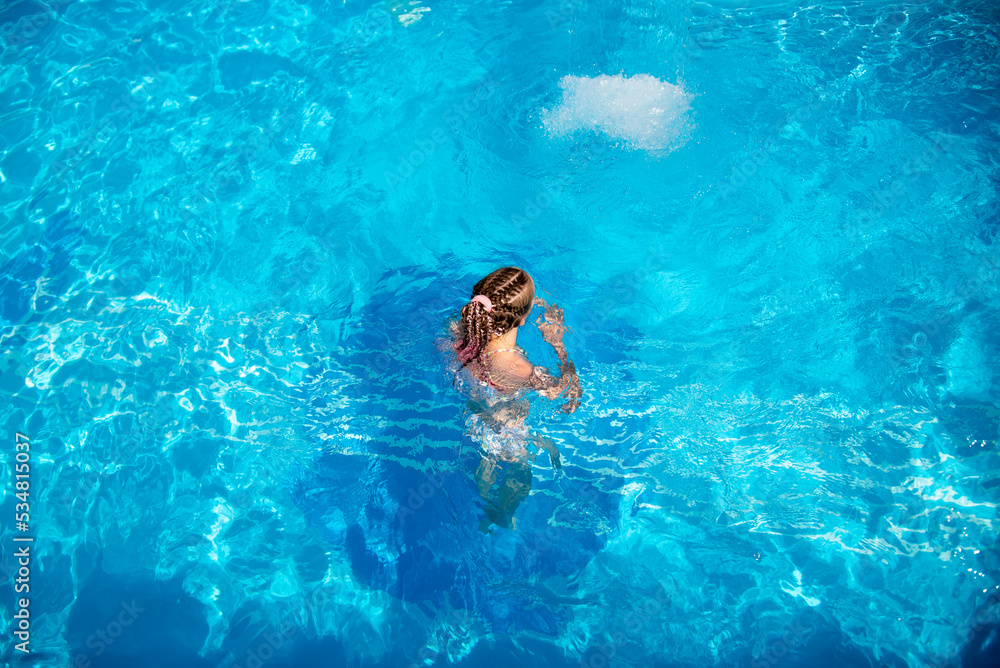 A teenage girl of 11-13 years old swims in a pool with blue water. She has african braids braided with zi-zi ribbons. Summer. Family vacation.