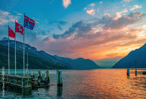 Stunning sunset over Lucerne Lake with burning sky reflected in the water. Flag of municipality Ingenbohl, National flag of Switzerland and flag of Canton of Schwyz, Brunnen, Switzerland
