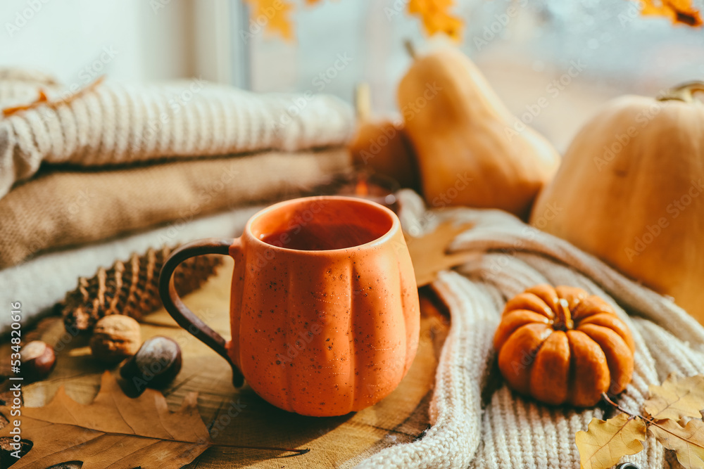 A cup of hot steaming tea and a pumpkin-shaped candle on the windowsill, autumn mood A cup of hot steaming tea and a pumpkin-shaped candle on the windowsill, autumn mood