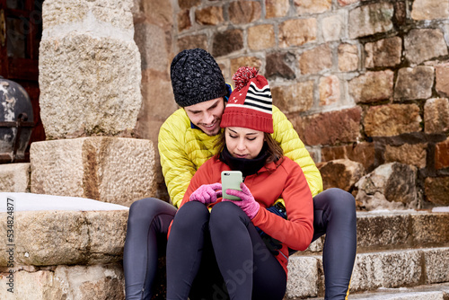 Couple in winter jackets and hats using cell phone sitting on stone steps.