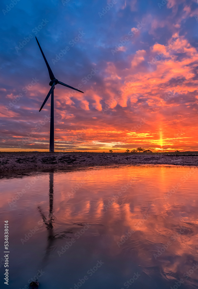 wind energy turbine silhouette with a frozen lake and reflection on ice and a colorful sunset sky on background