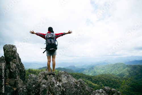 Young person hiking female standing on top rock  Backpack woman looking at beautiful mountain valley at sunlight in summer  Landscape with sport girl  high hills  forest  sky. Travel and tourism.