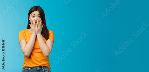 Shocked speechless stunned asian girl stare camera cover mouth palms, raise eyebrows gasping astonished, feel pitty sorry hearing bad news, stand surprised blue background wear yellow t-shirt photo