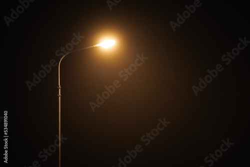 Naklejka One night lamppost shines with faint mysterious yellow light through evening fog. Streetlight shine at quiet city night, magic atmospheric light in mystical darkness, copy space