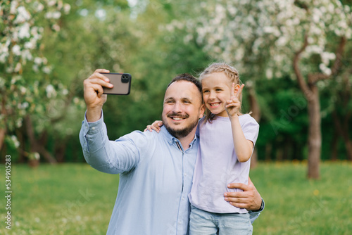 Close-up portrait of a happy family of two. Smiling dad and six-year-old daughter take a selfie with a smartphone in a green spring or summer park.