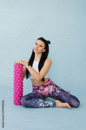 Fitness a woman in sportswear with a massage roller or a roll in her hands smiles while sitting on a blue isolated background. Fitness trainer with items for myofascial release