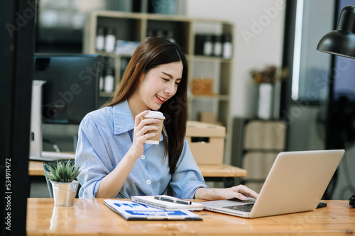 Confident beautiful Asian businesswoman typing laptop computer and digital tablet while holding coffee at office