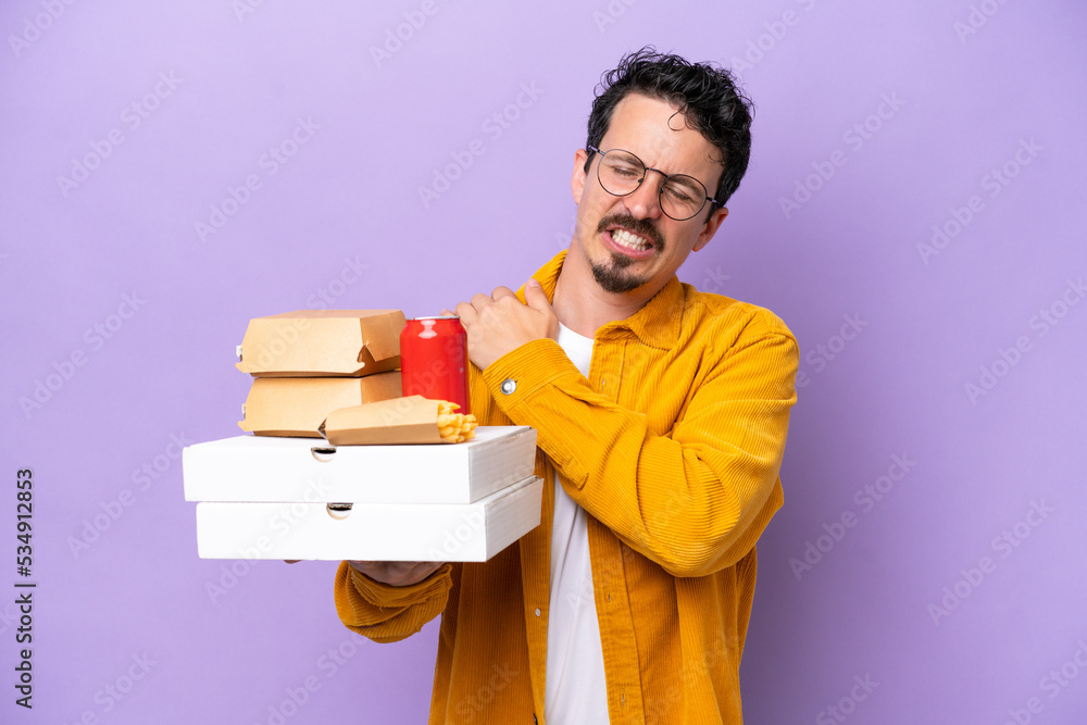 Young caucasian man holding fast food isolated on purple background suffering from pain in shoulder for having made an effort Young caucasian man holding fast food isolated on purple background suffering from pain in shoulder for having made an effort