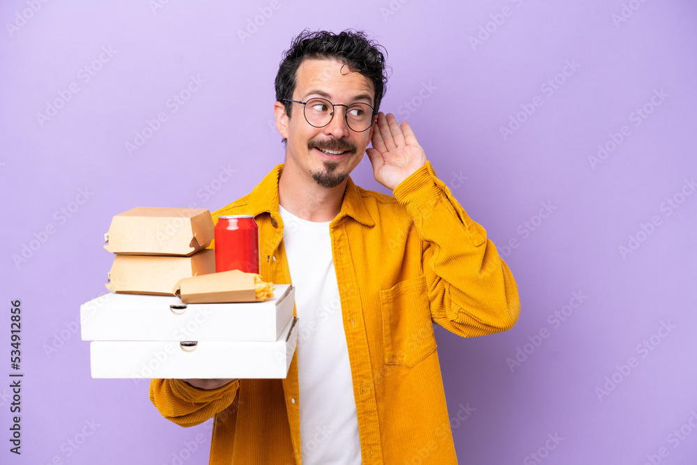 Young caucasian man holding fast food isolated on purple background listening to something by putting hand on the ear Young caucasian man holding fast food isolated on purple background listening to something by putting hand on the ear