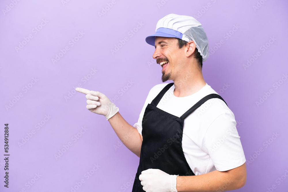 Fishmonger man wearing an apron isolated on purple background pointing finger to the side and presenting a product