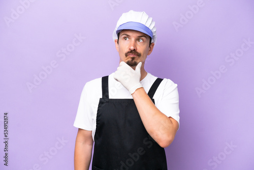 Fishmonger man wearing an apron isolated on purple background having doubts