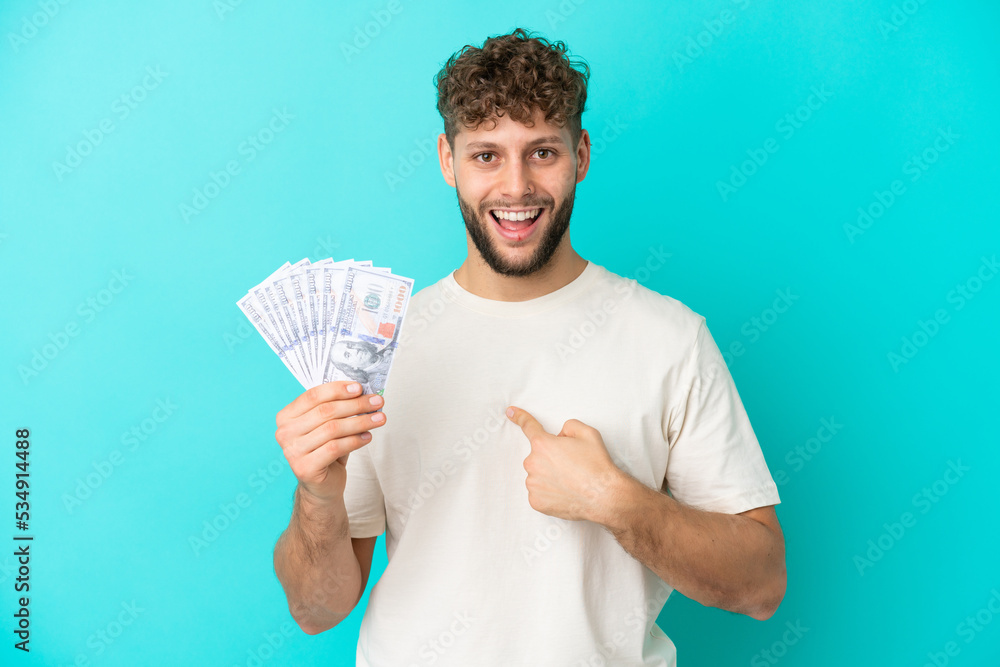 Young caucasian man taking a lot of money isolated on blue background with surprise facial expression Young caucasian man taking a lot of money isolated on blue background with surprise facial expression
