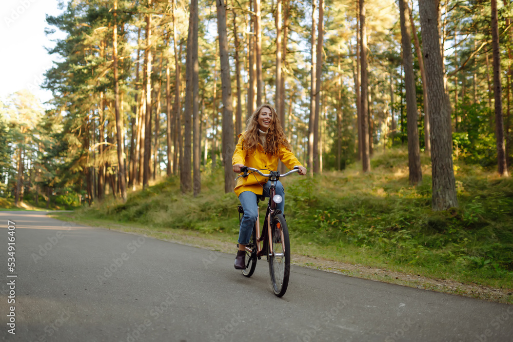 Fototapeta premium Beautiful happy woman in yellow coat riding bicycle in autumn park. Autumn fashion. Lifestyle. Relax, nature concept.