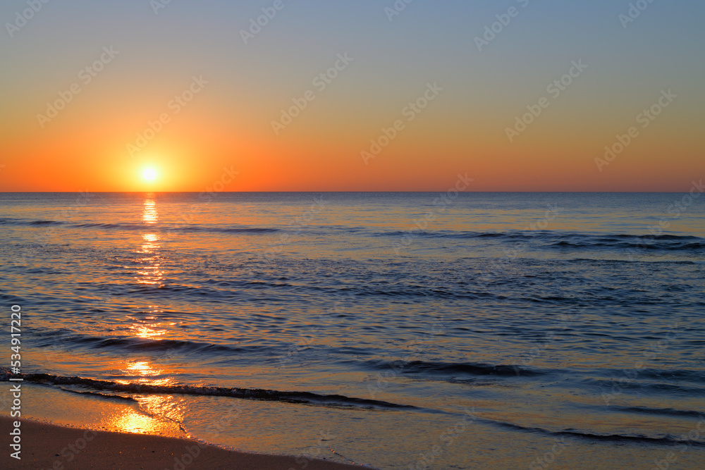 Die Sonne geht mit Orange Tönen über dem blauem Meer unter und leichte Wellen schwappen an den Strand.