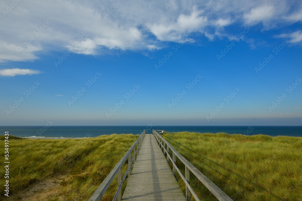 Ein Brücke aus Holz führt zum Schutz der Küstenlandschaft über eine Grasbewachsene Sanddüne zum Strand. Das Meer leuchtet Blau und am blauem Himmel hängen leichte weiße Wolken.