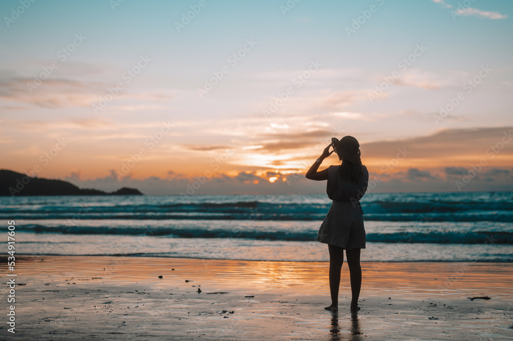 Silhouette of happy woman in her hat is back and admire the sunset over the sea on the beach
