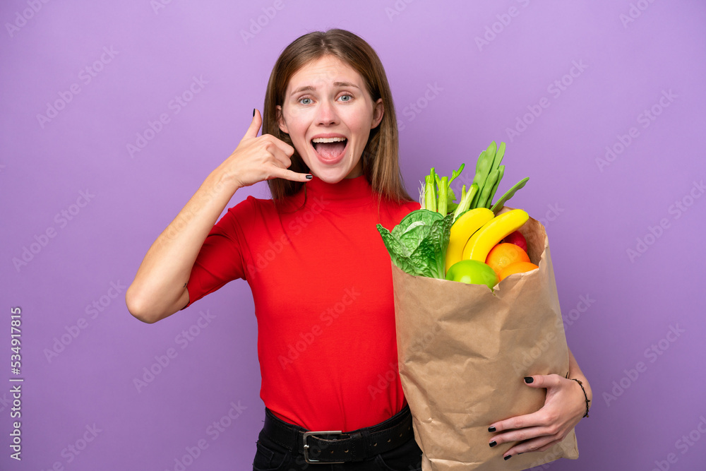 Young English woman holding a grocery shopping bag isolated on purple background making phone gesture. Call me back sign Young English woman holding a grocery shopping bag isolated on purple background making phone gesture. Call me back sign