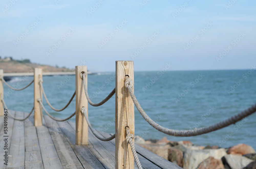 Fototapeta premium Wooden platform on the seashore. Fence made of old marine rope. Rope knot. Wooden poles. Yacht mooring.