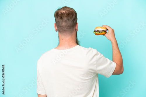 Redhead man with long beard holding a burger isolated on blue background in back position