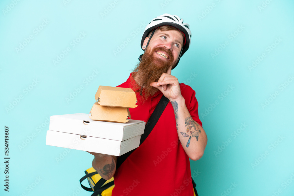 Delivery man holding pizzas and burgers isolated on blue background thinking an idea while looking up Delivery man holding pizzas and burgers isolated on blue background thinking an idea while looking up
