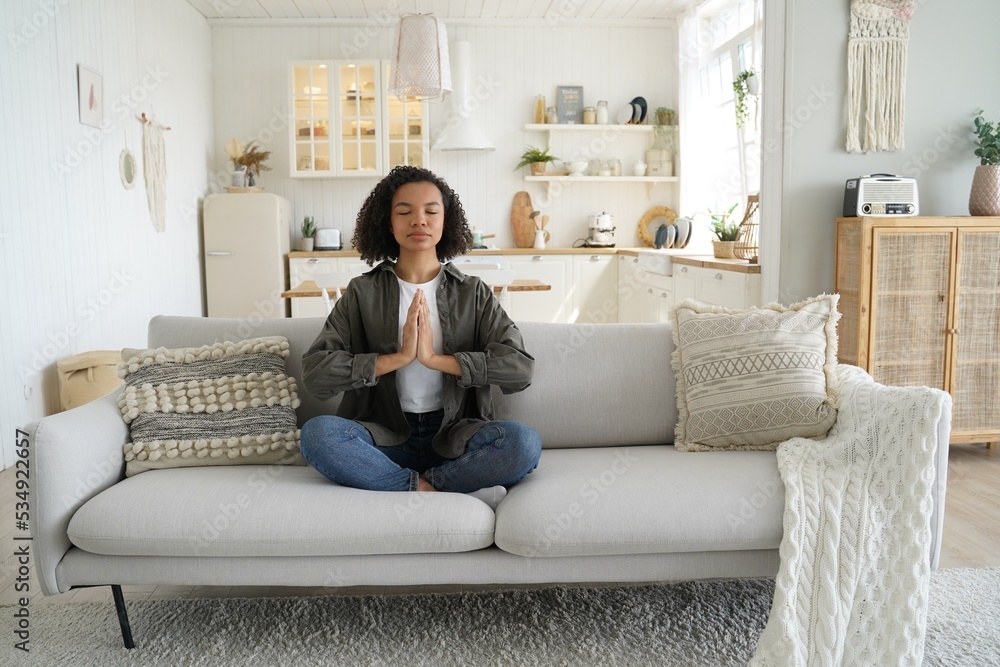 Mixed race girl practices yoga, meditation at home sitting in lotus pose on couch. Healthy lifestyle Mixed race girl practices yoga, meditation at home sitting in lotus pose on couch. Healthy lifestyle