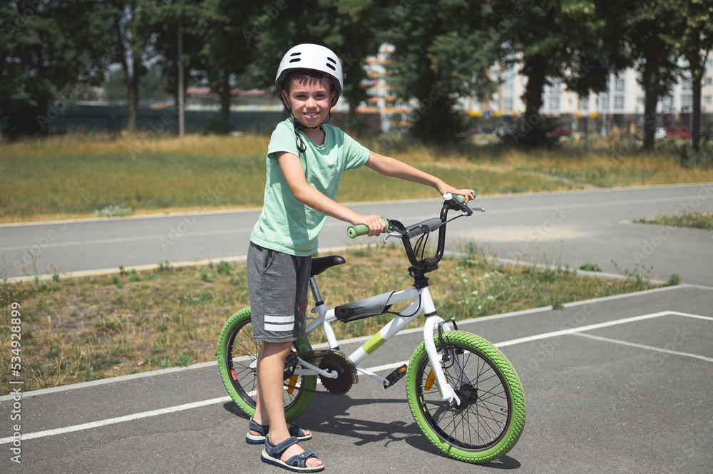 Charming active Caucasian child boy, in protective helmet, looking at camera, standing near his bicycle on the asphalt road outdoor. Physical activity. Active lifestyle. Equilibre and balance. Sport