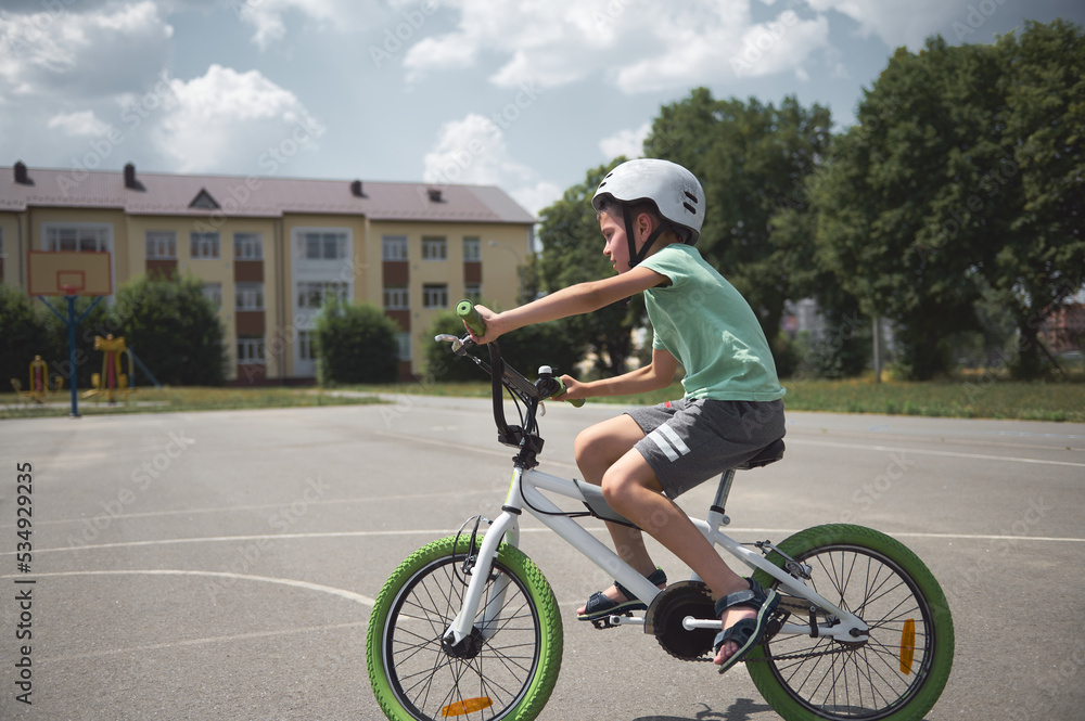 Obraz premium Side full-length of a sporty child boy in protective sports helmet, riding a bicycle in the city asphalt road. Active elementary age kid learning extreme cycling and tricks on his bike. Equilibre