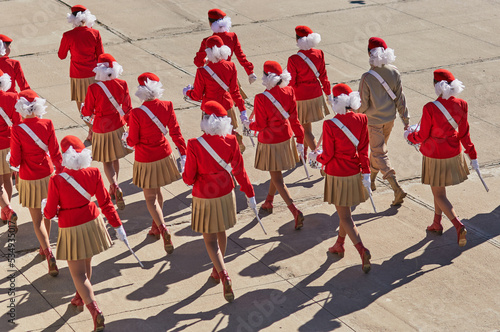 A group of girls with drums march in formation at the parade. The drummers are dressed in red uniforms. Sunny day. Side view from above. Identical white bows on the head
