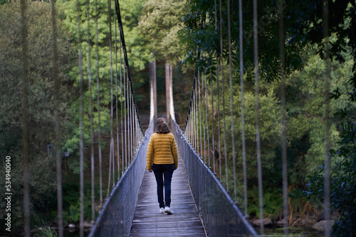 Close view of a woman with a yellow anorak walking on the suspension bridge with metal fence over the Miño river in Spain in an environment of native trees. 